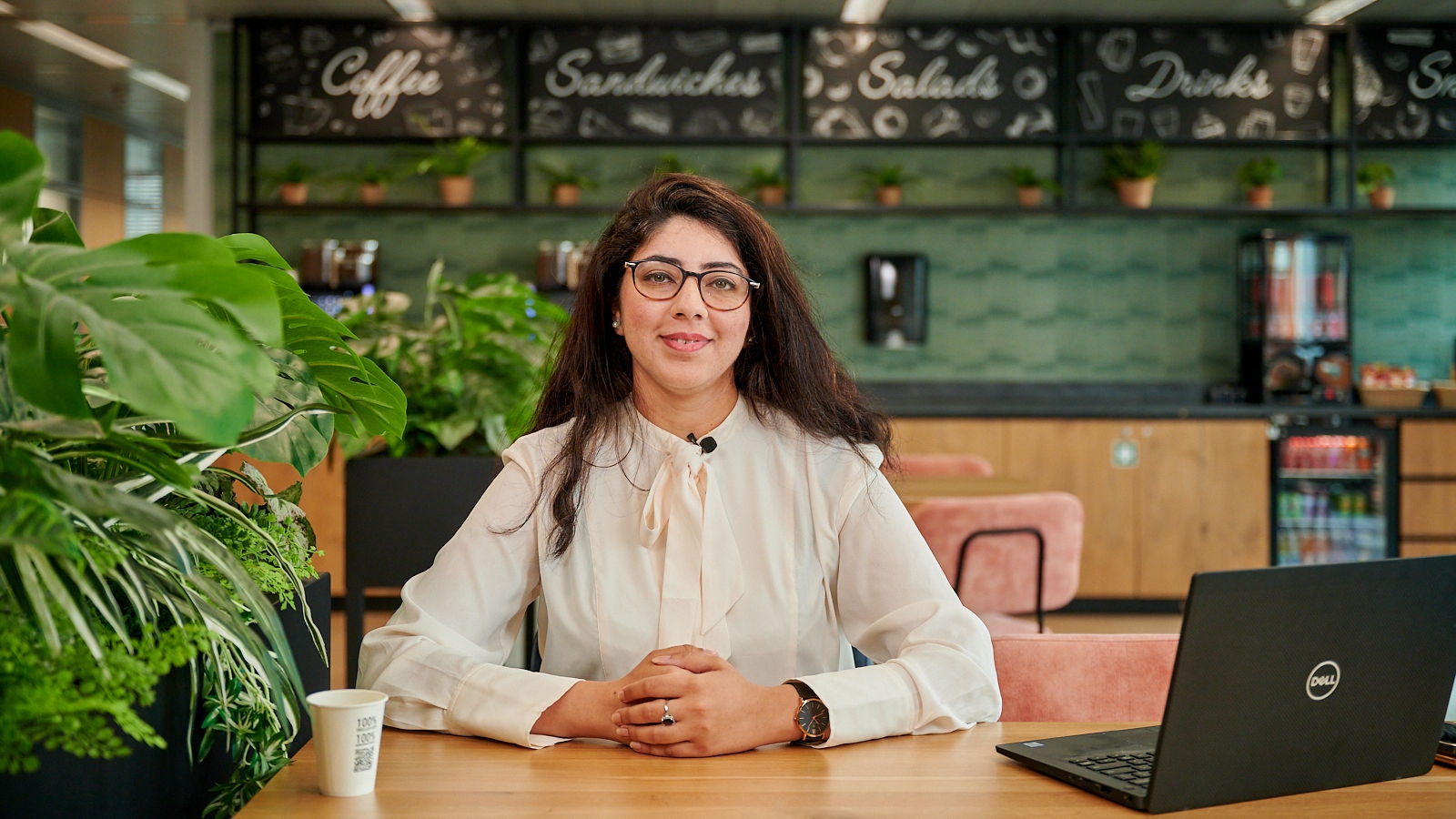 Girl with dark hair and glasses smiling at the camera, seated near the coffee corner with her laptop and cup of coffee.