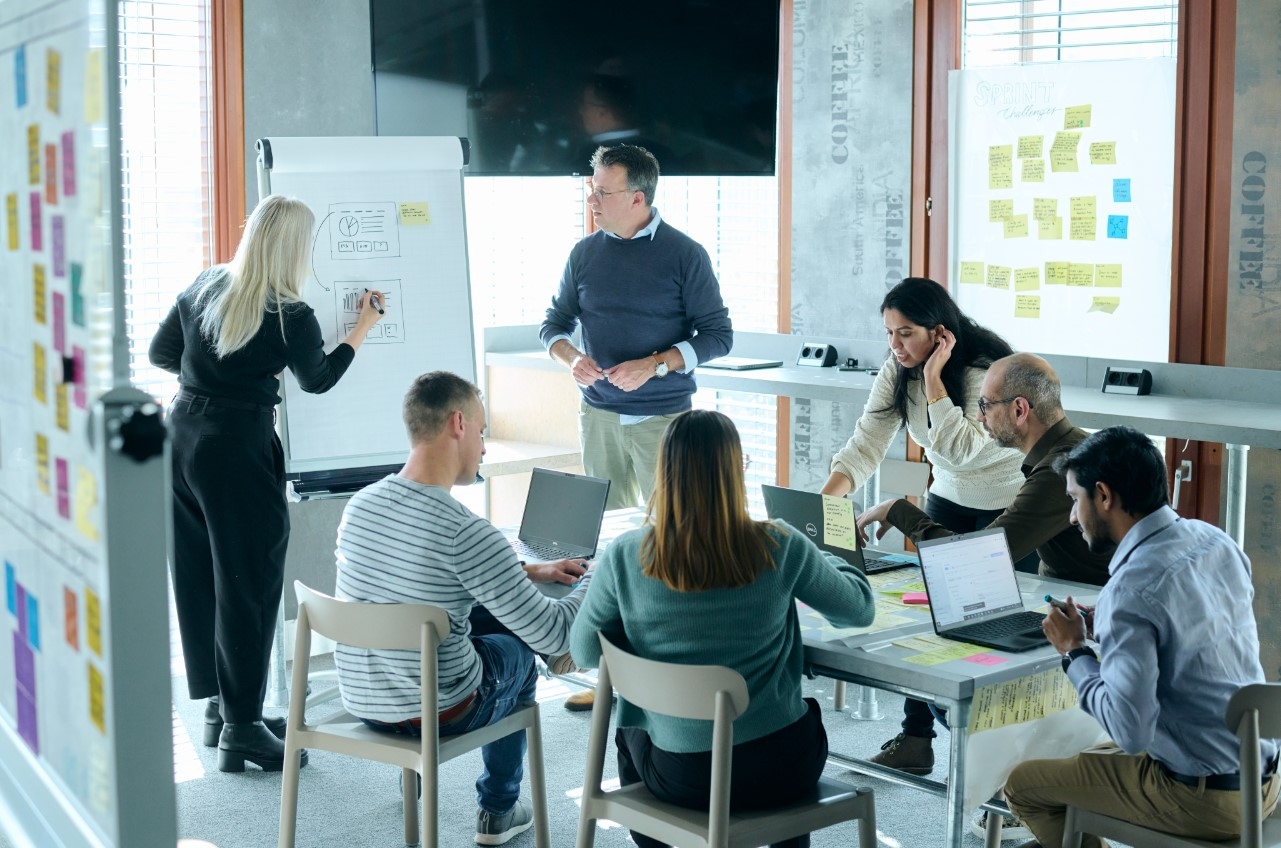 Six people in a meeting room collaborating on a project, with some using a laptop and others writing on a whiteboard, all working together as a team.