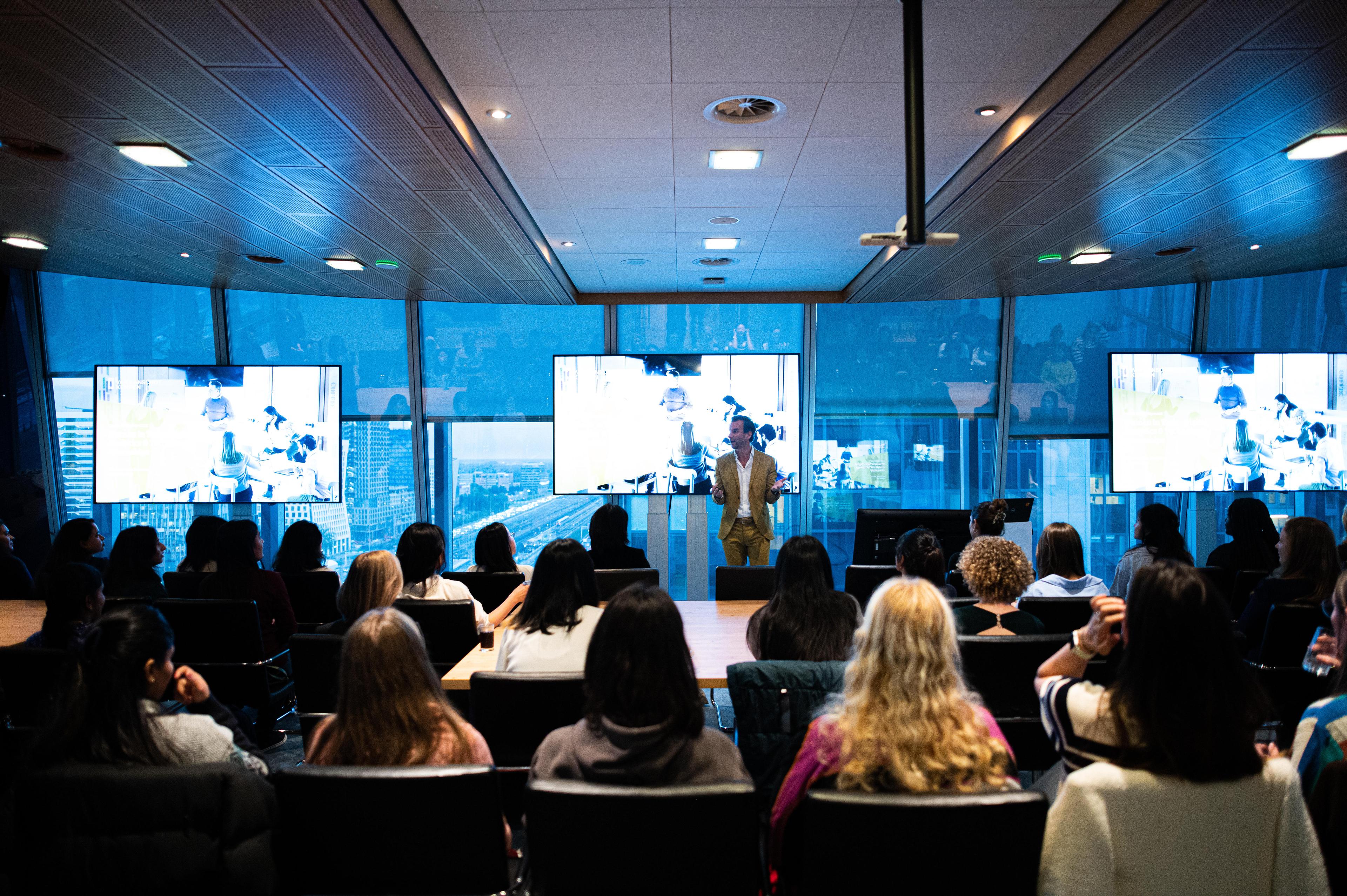 View of the Zonnezaal room filled with attendees seated and listening attentively to a presenter at the front, delivering a presentation.